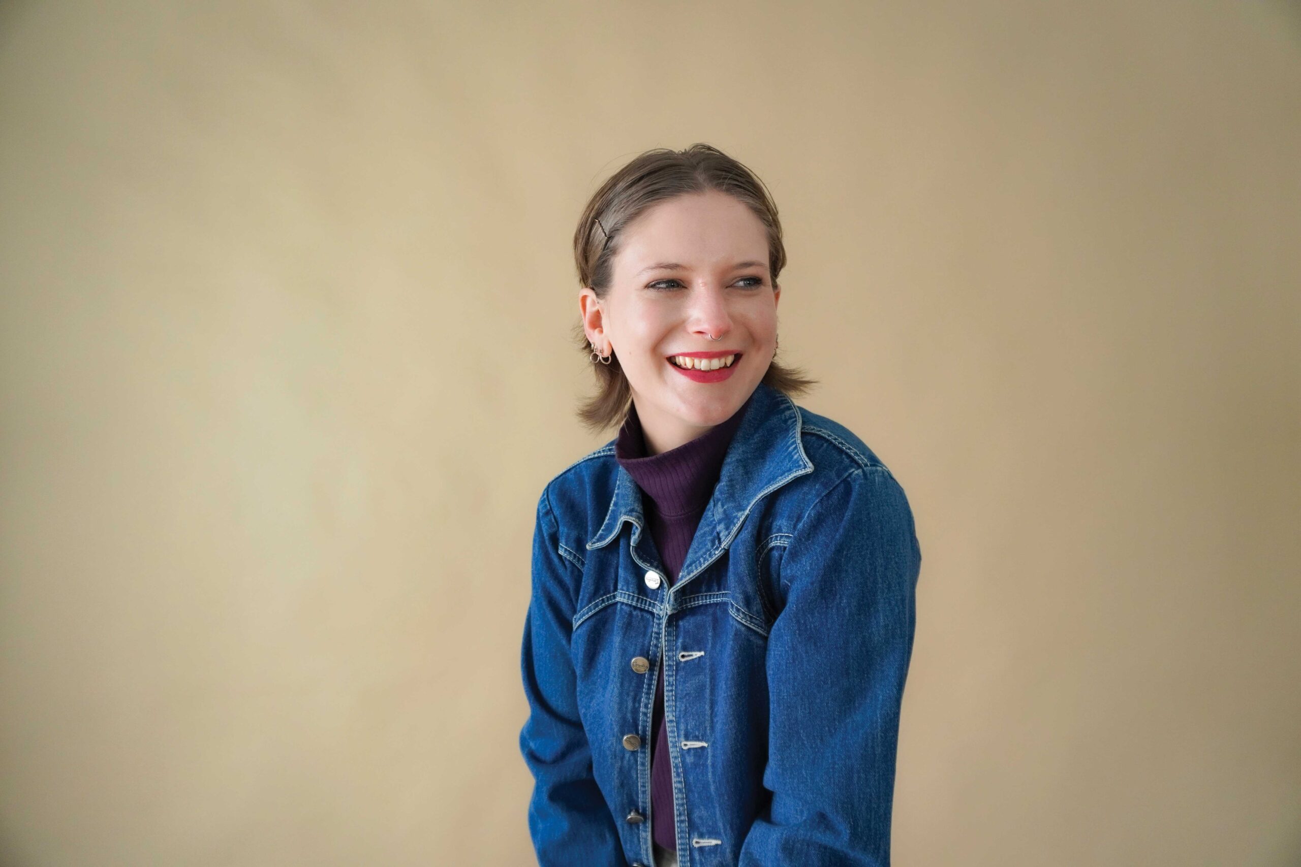 Smiling woman in a denim jacket and purple turtleneck against a beige background