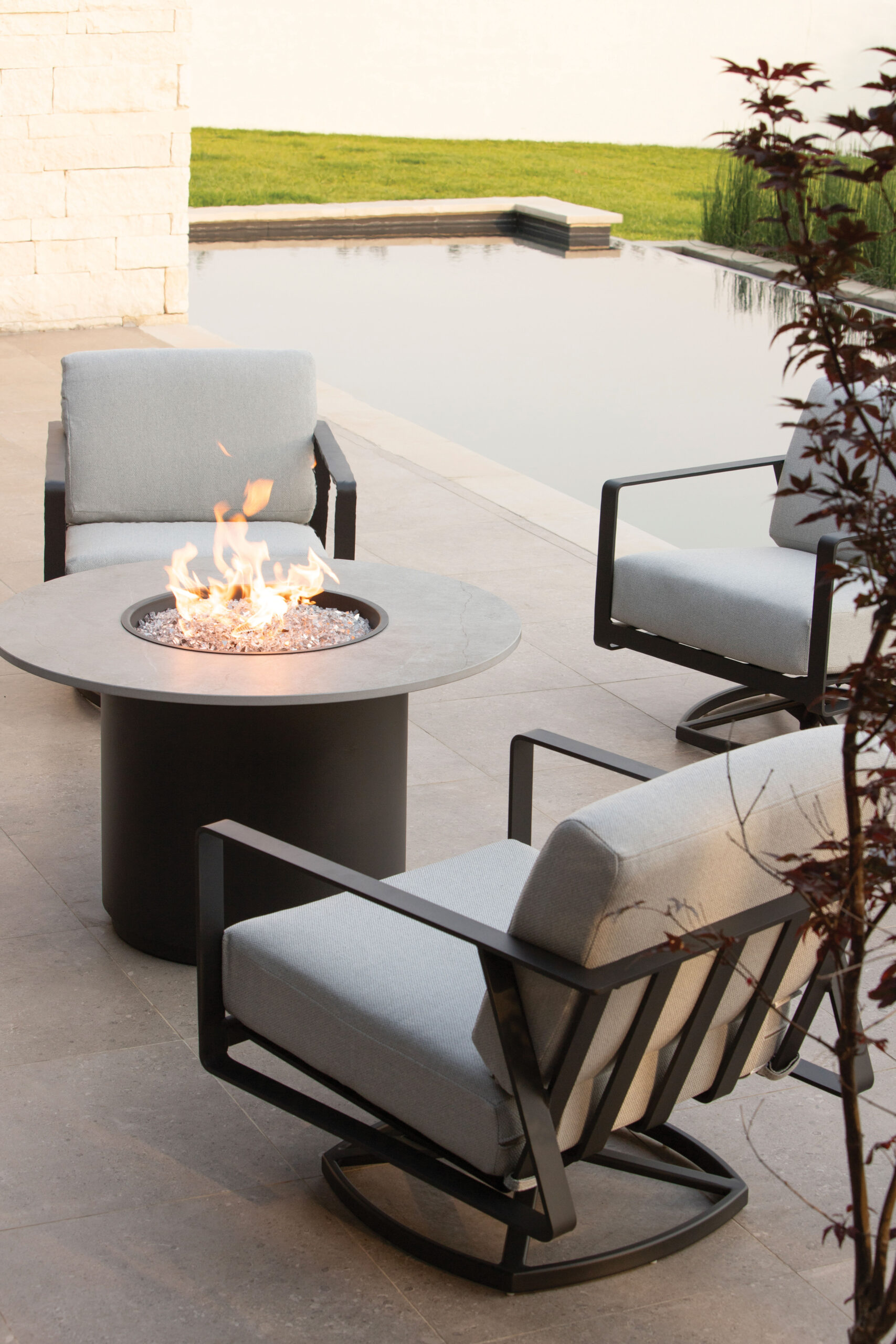 Modern dark-framed patio chairs with gray cushions围 a round fire pit table beside a reflecting pool
